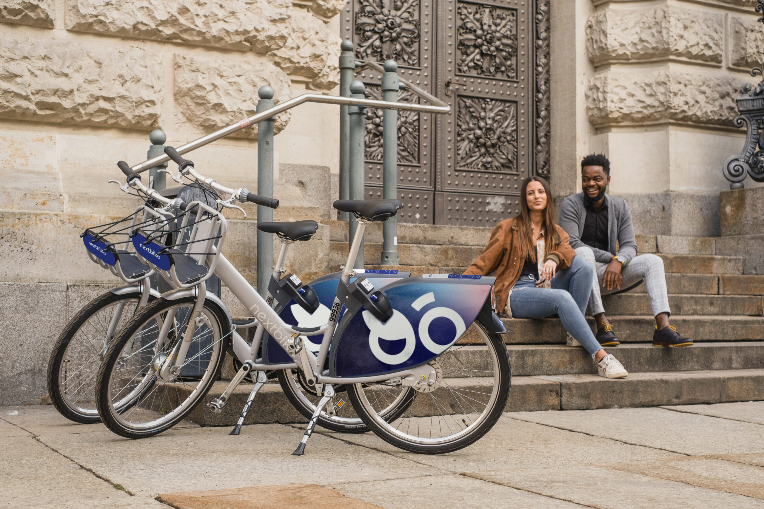 Next bike photo of a woman and a man sitting on steps waching 2 parked nexbikes -Micro Mobility